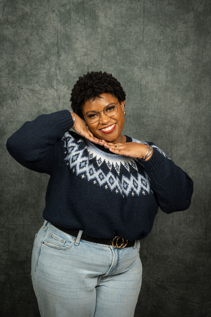 Smiling person in glasses and a retro sweater, posing for an 80's themed photoshoot.