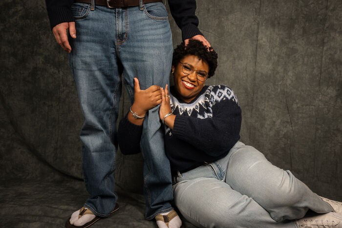 Couple in 80s-themed photoshoot, man wears jeans, woman sits smiling in a sweater, capturing an awkward anniversary moment.