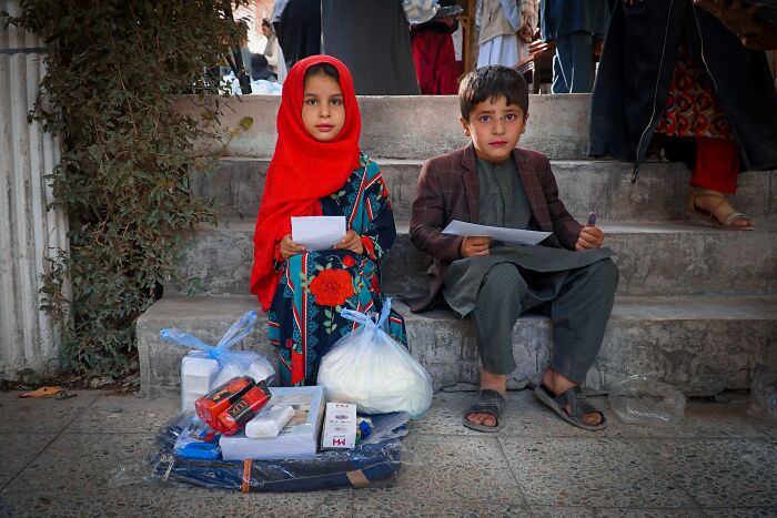 Two Afghan children sitting on steps holding papers with aid supplies, illustrating life while traveling in Afghanistan.