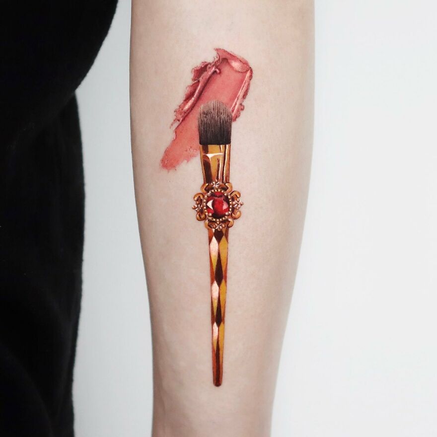 Golden tattoo of a makeup brush and lipstick smear, crafted with precision by Manhattan-based artist.