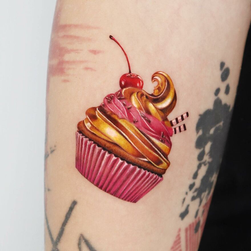 Golden tattoo of a cupcake with cherry, created by skilled Manhattan-based artist.