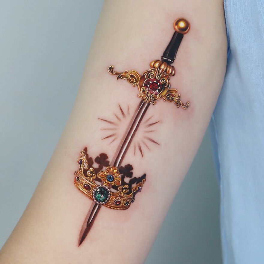 Golden tattoo of a jeweled sword and crown on forearm, designed by Manhattan artist.