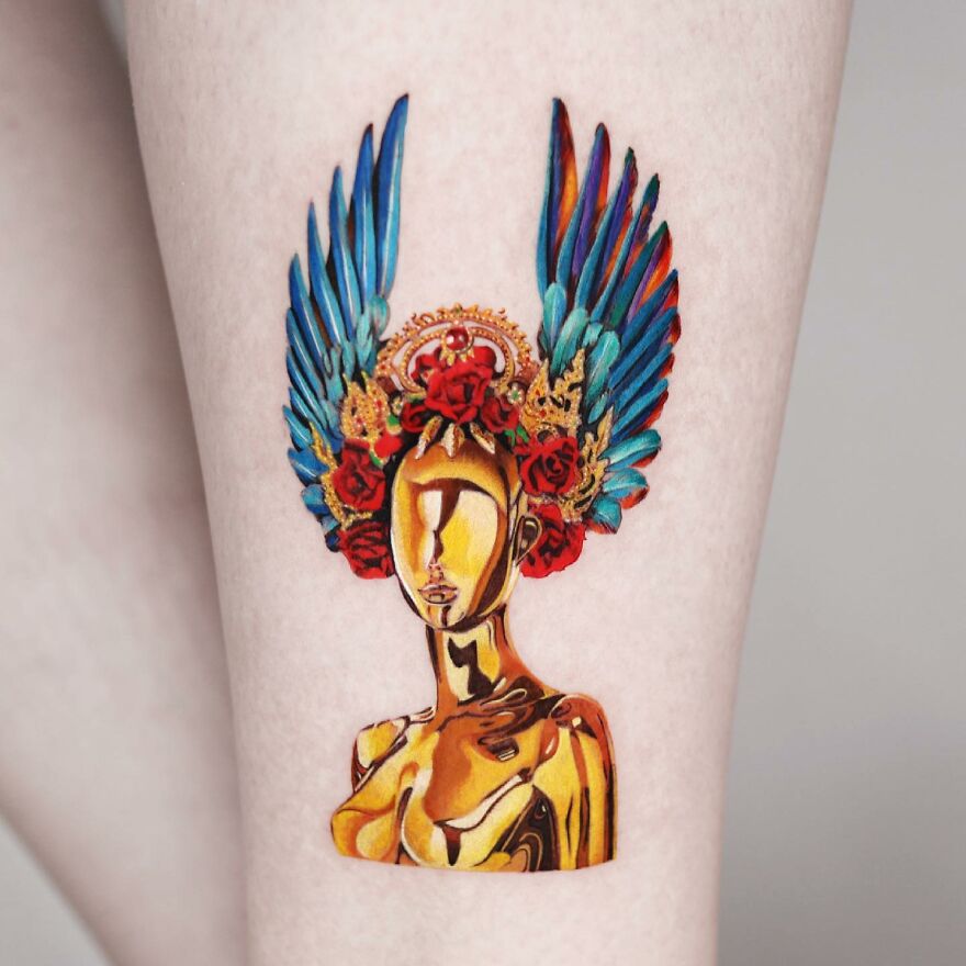 Golden tattoo of a stylized figure with blue feathered wings and red roses on the arm, created by a Manhattan-based artist.