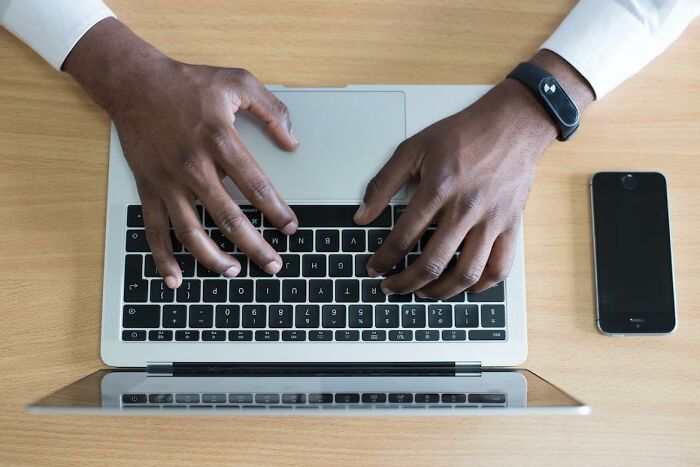 Person wearing fitness tracker typing on laptop keyboard with smartphone on wooden desk, illustrating things everyone does.