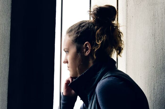Young woman with a bun wearing a dark jacket, looking thoughtfully out a window in a quiet, dimly lit space.