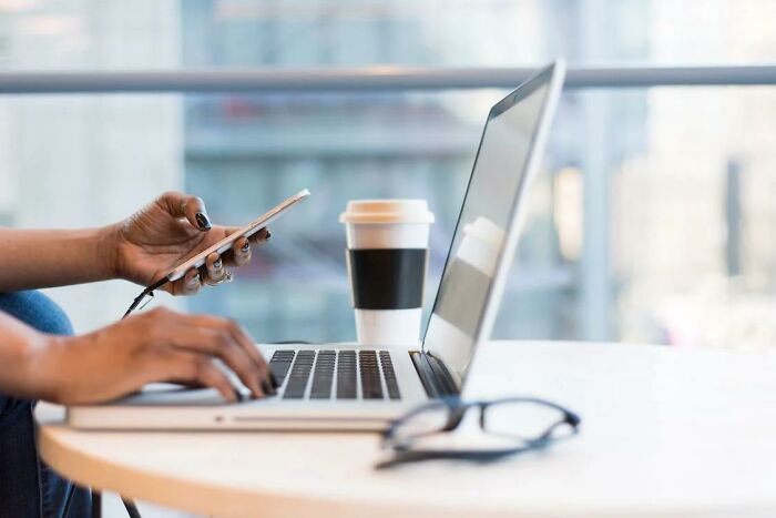 Person using smartphone and laptop at a table with coffee cup, illustrating concept of criminally overpriced items discussion.