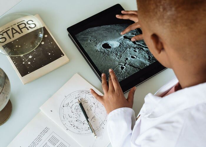 Child exploring moon crater image on tablet with astronomy books and star charts, highlighting criminally overpriced interests.