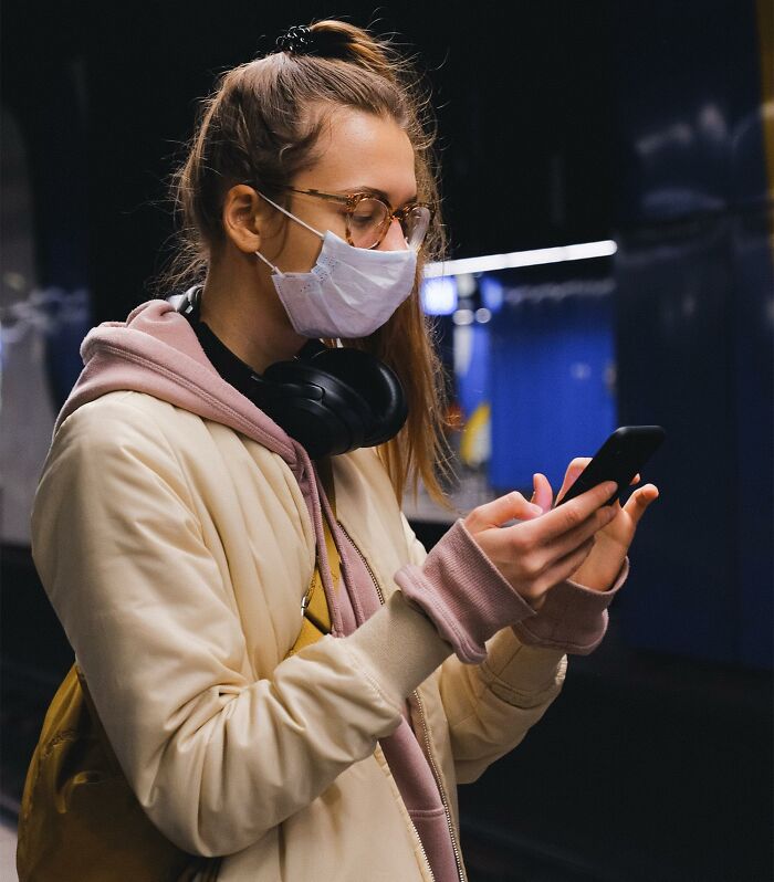 Young woman wearing a face mask and glasses, using smartphone while waiting at a subway station, illustrating life-saving tips.