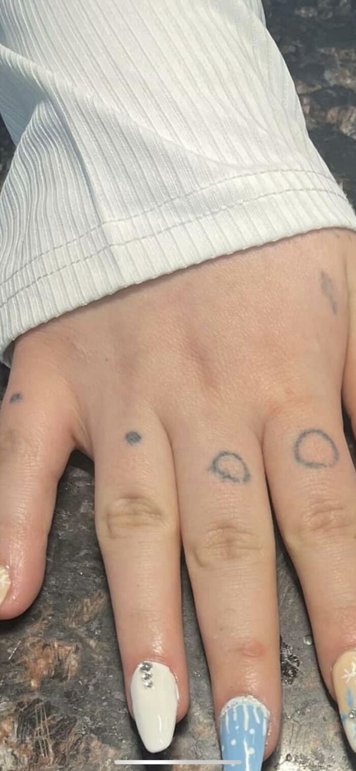 Hand with small, humorous tattoos on fingers alongside white and blue painted nails.