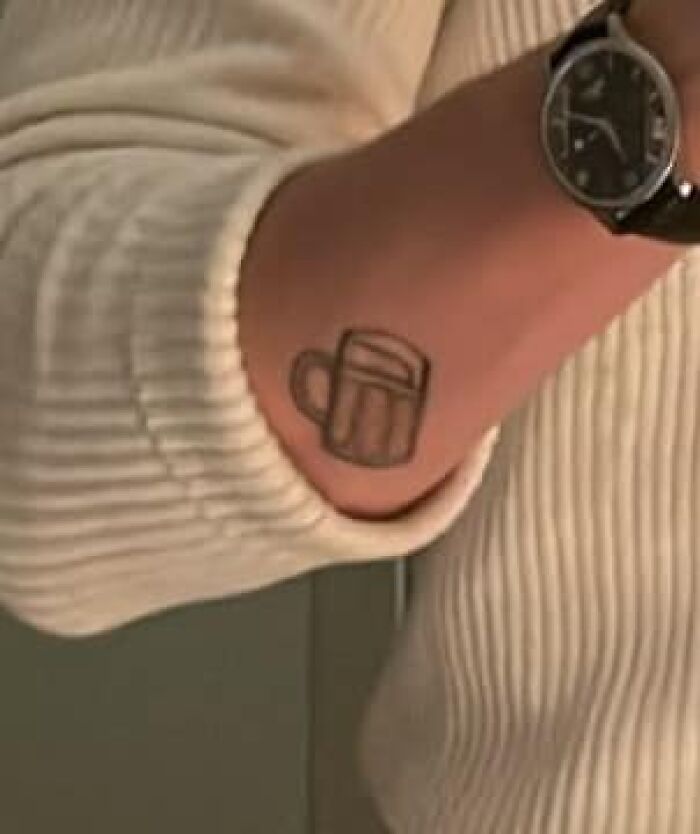A tattoo of a simple beer mug on a person’s forearm, next to a watch, showcasing a funny bad tattoo.