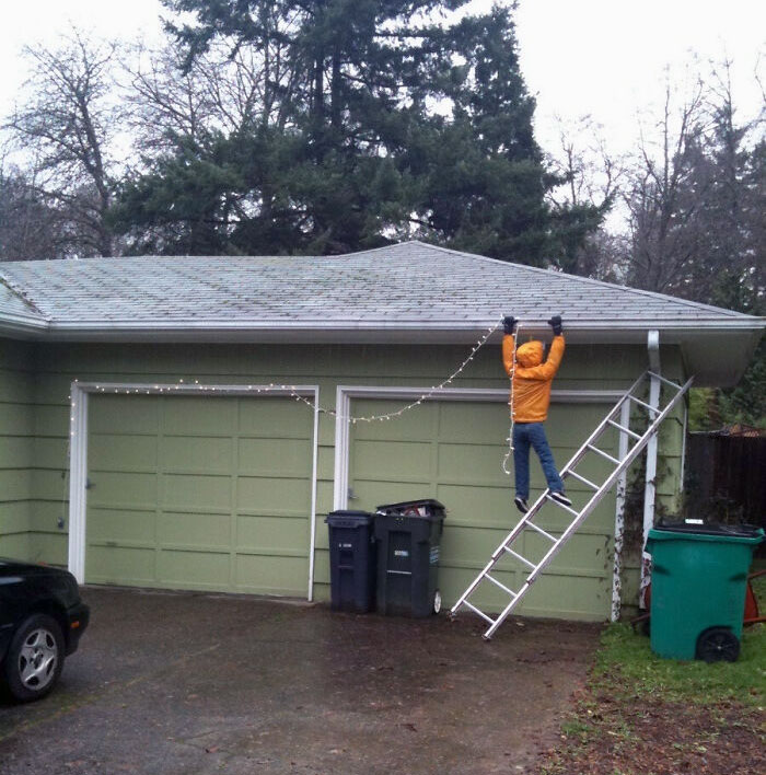 Person in a yellow jacket humorously hanging Christmas lights on a garage roof using a ladder.