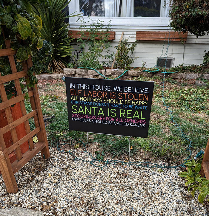 Humorous Christmas sign with playful messages in a garden setting.