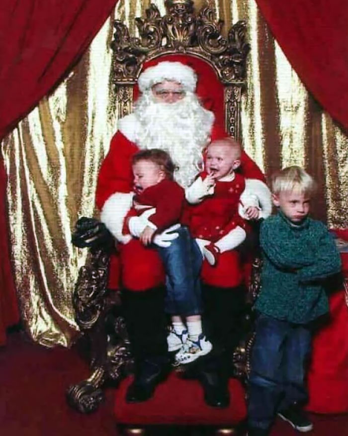 Santa posing with three kids, two crying on his lap and one pouting beside him, adding humor to a Christmas scene.