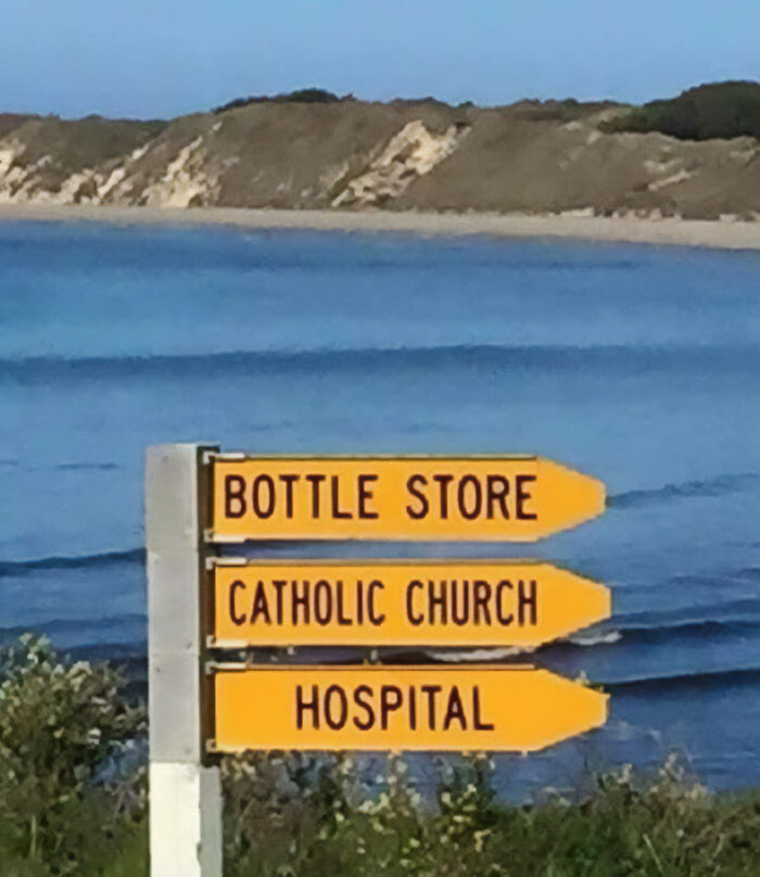 Yellow directional signs for bottle store, catholic church, and hospital by the sea, highlighting unique Ireland culture differences.