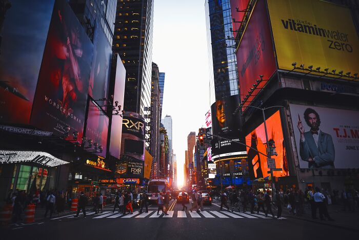 Crowded city street at sunset with pedestrians crossing and brightly lit billboards, capturing urban life and community fears.