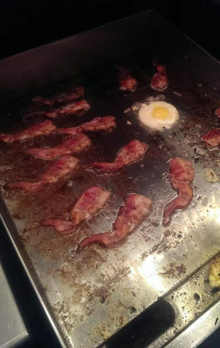 Bacon strips and a fried egg cooking on a greasy griddle, featured in a group rating others dishes online.