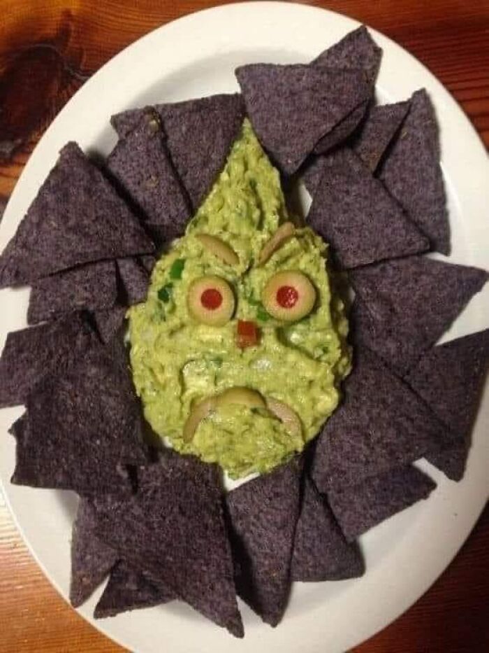 Guacamole shaped like an angry face surrounded by blue corn tortilla chips on a white plate in a food rating group.