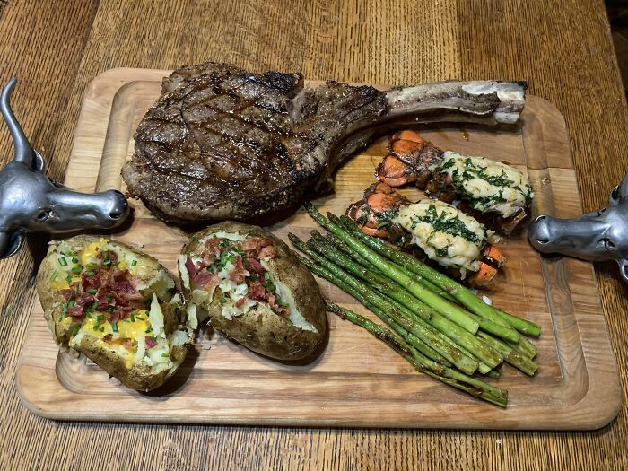 Steak, loaded baked potatoes, grilled asparagus, and stuffed lobster tails served on a wooden board in a food rating group.