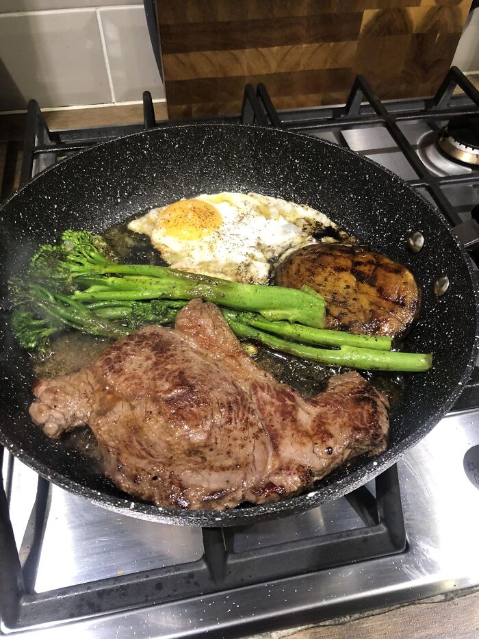 Pan frying steak, broccolini, egg, and mushroom on a stovetop in a cooking pan for dish rating in Facebook group.