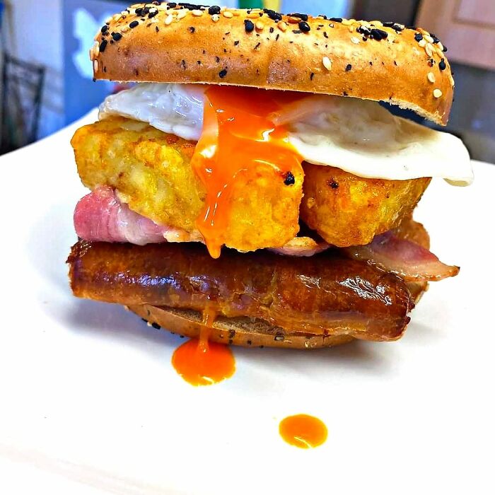 Breakfast sandwich with sausage, bacon, fried egg, hash browns, and spicy sauce on a seeded bagel in food rating group.