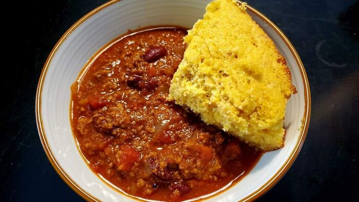 Bowl of chili with ground beef and beans served with a golden cornbread slice in a food rating Facebook group.