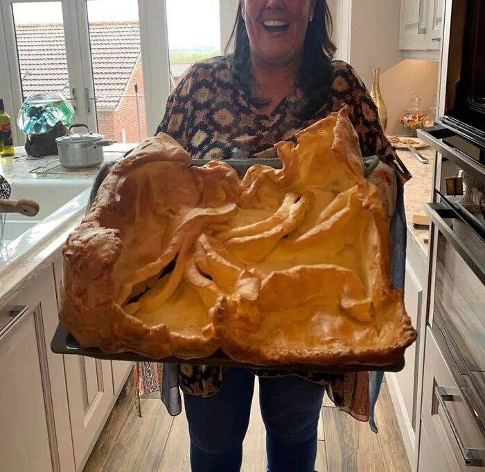 Woman holding an oversized, unevenly baked pie in a kitchen, featured in a Facebook group rating others' dishes.