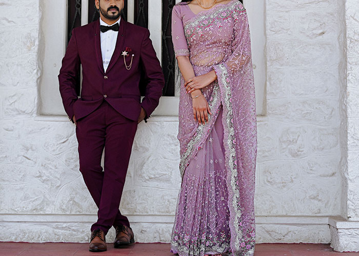 Man in a burgundy suit and woman in a purple saree standing against a white textured wall, illustrating men’s annoying experiences.