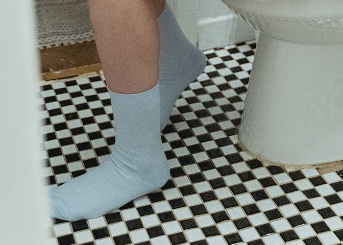 Person wearing light blue socks standing on black and white tiled bathroom floor near a toilet, illustrating men’s everyday annoyances.