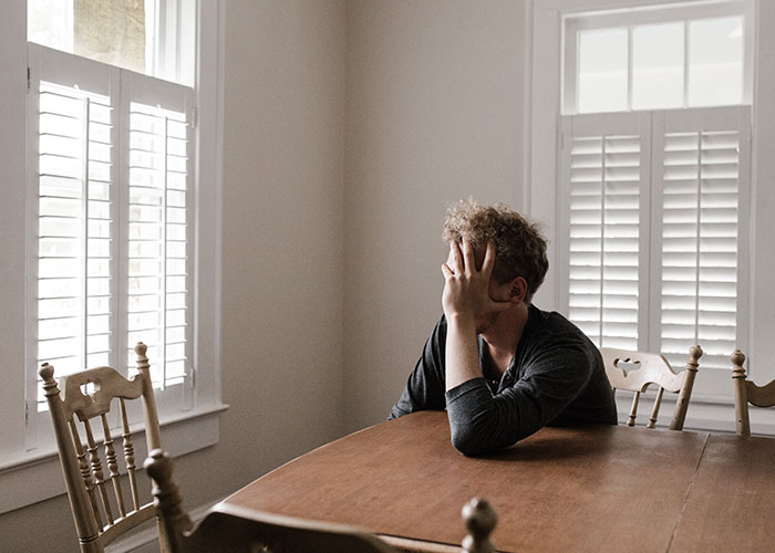 A man sits alone at a wooden table, covering his face, illustrating frustration from annoying experiences men face due to gender.