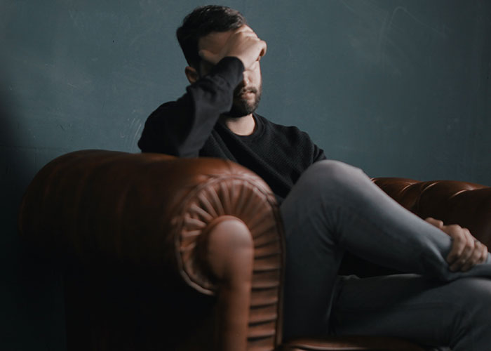 Man sitting on a brown leather couch, holding his forehead, expressing frustration with challenges men face because of their gender.