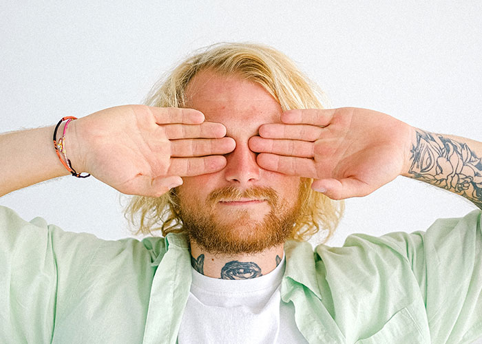 Young man with tattoos covering eyes with hands, illustrating annoying things men experience because of their gender.