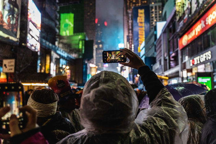 Crowd capturing city lights and celebrations on New Year's Eve with smartphones, wearing raincoats for protection.