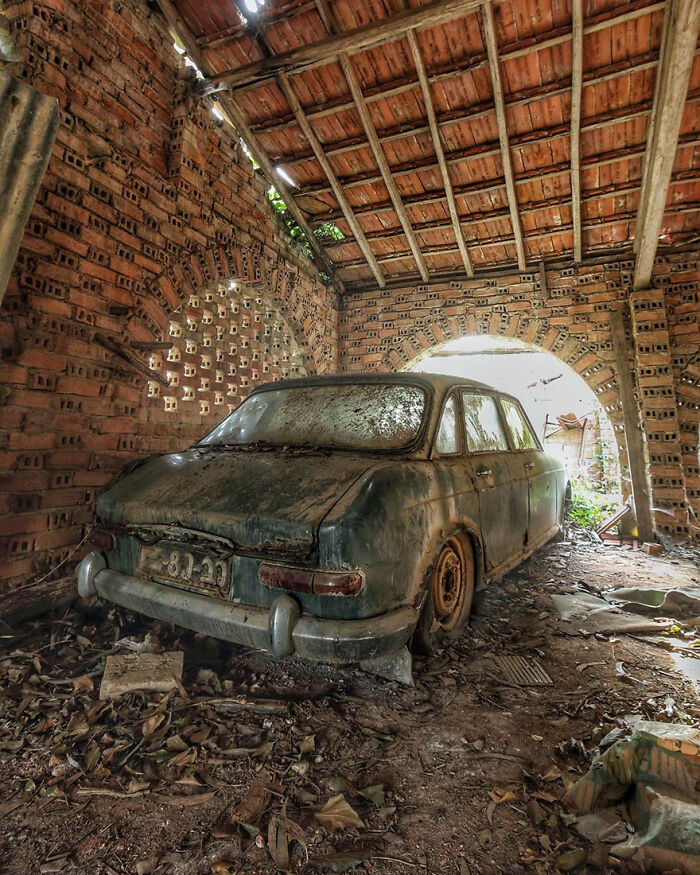 Abandoned classic car covered in dust inside a decaying brick factory with debris and natural light filtering in.