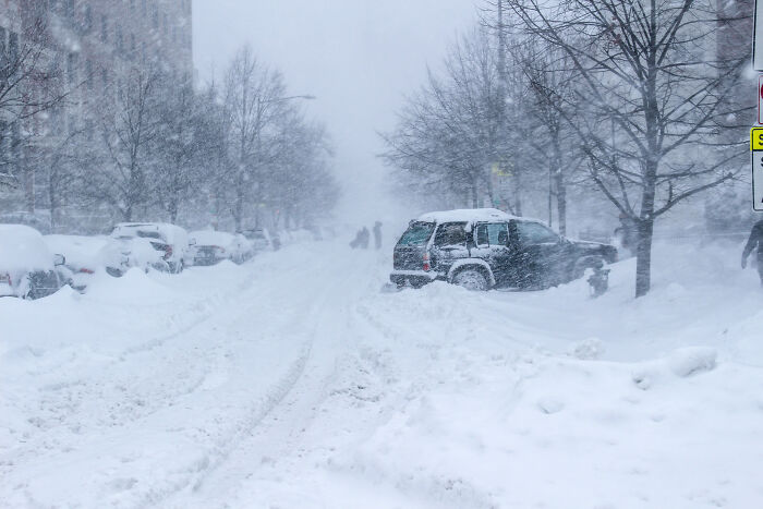 Snow-covered street with parked cars and people walking in a heavy snowstorm, highlighting noticeable everyday challenges.