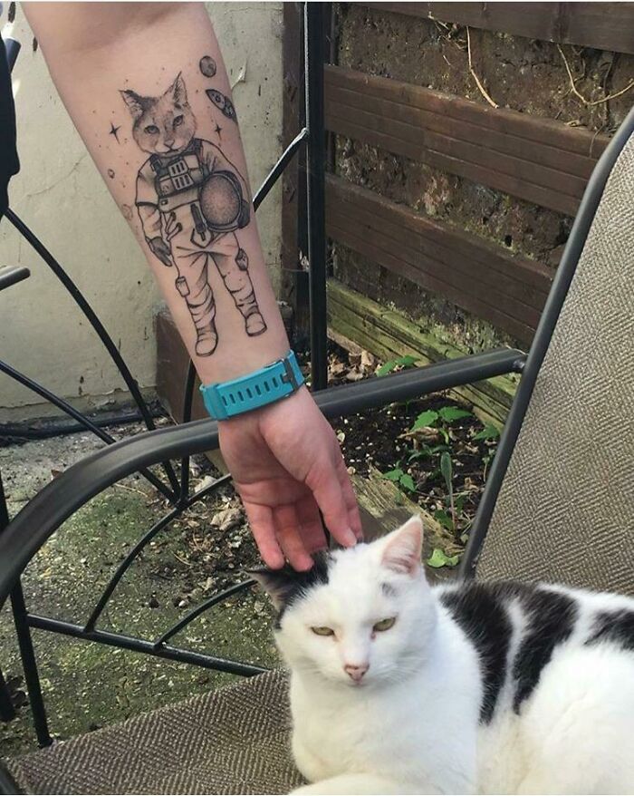 Beautiful tattoo of a cat astronaut on an arm, with a white cat being petted nearby.