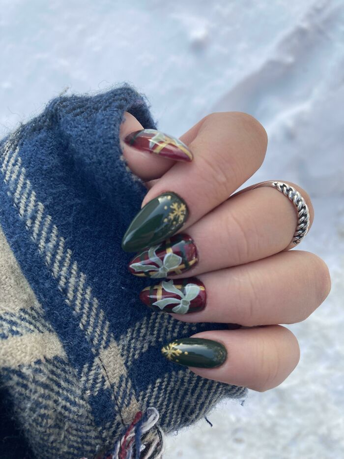 Hand with winter-themed nail art ideas featuring green polish, gold snowflakes, and red plaid patterns on a blue plaid sleeve.