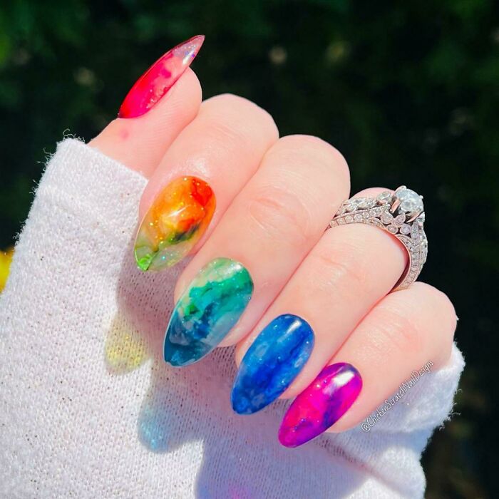 Colorful marble nail art with rainbow design on fingers, featuring a diamond ring and a white sleeve.