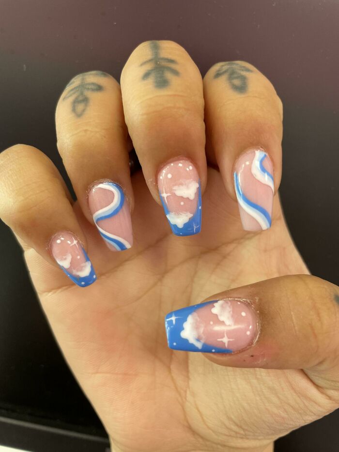 Hand with nail art featuring blue and white cloud and swirl designs on a n**e base, showcasing creative nail art ideas.