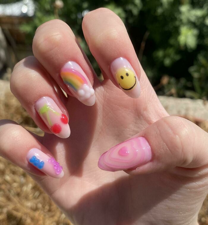 Hand with colorful nail art including a rainbow, smiley face, cherries, bears, and heart designs in bright pastel colors.