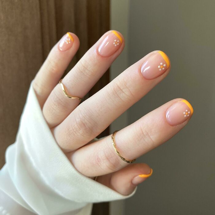 Close-up of hand with nail art featuring orange French tips and small white floral designs on natural nails.
