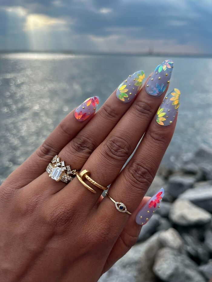 Hand with colorful floral nail art designs and gold rings held up against a blurred ocean background showcasing nail art ideas.
