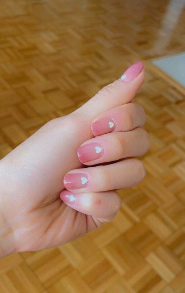 Hand showing nail art with pink polish and small white heart designs on each nail against wooden floor background.