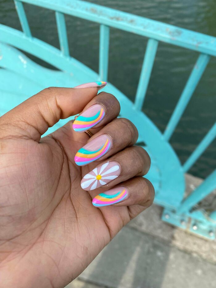 Hand with colorful nail art featuring rainbow swirls and a white daisy design by a turquoise railing over water.