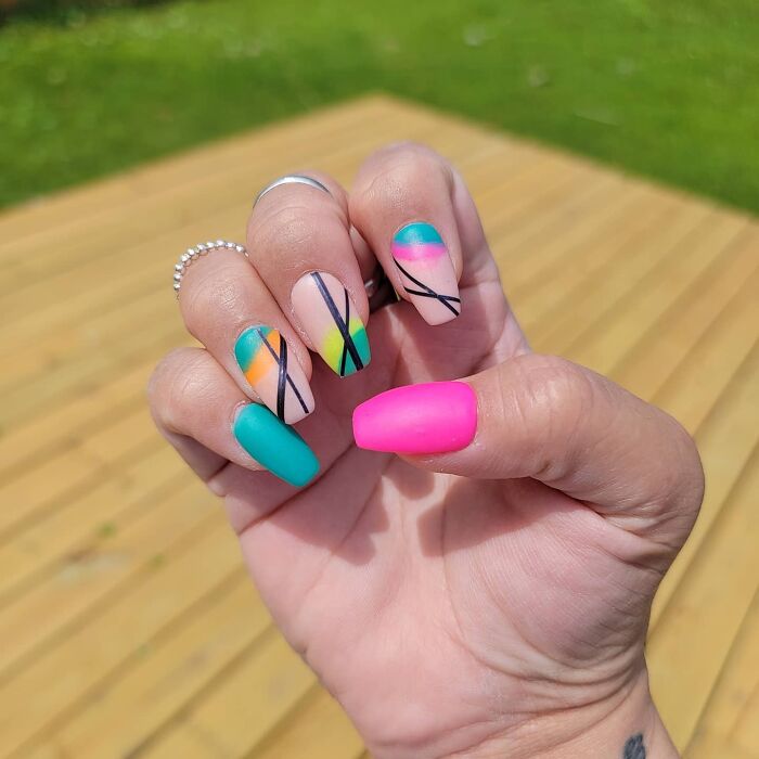Hand showing colorful nail art designs with geometric patterns and bright solid colors outdoors on wooden deck.