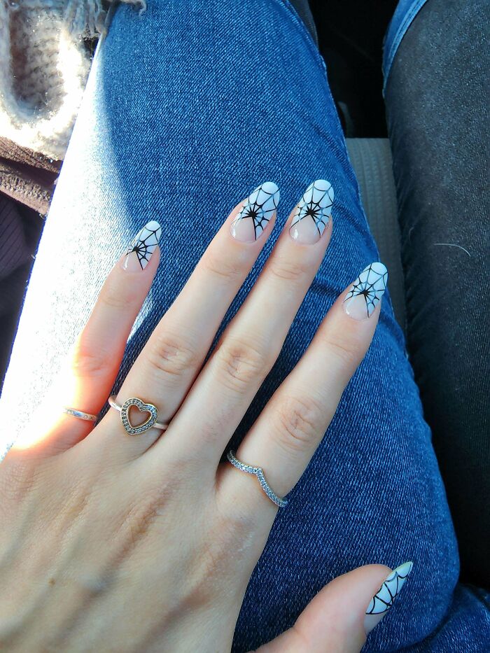 Hand with black and white spider web nail art design, wearing silver rings on blue denim background.