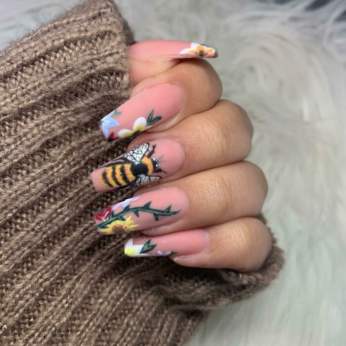 Hand with bee and floral nail art designs on long nails, showcasing creative nail art ideas on a neutral background.