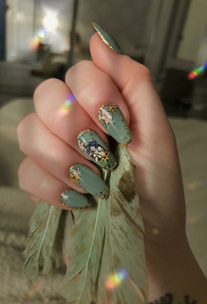 Hand showcasing green nail art ideas with floral and character designs, holding light-colored feathers indoors.
