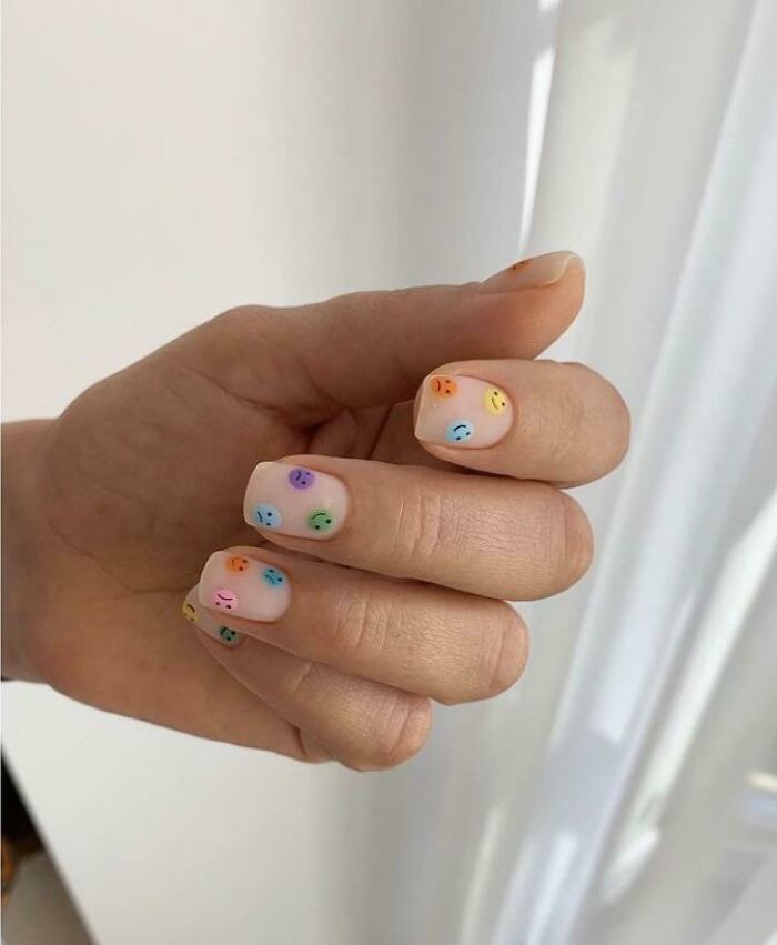 Hand with colorful smiley face nail art designs on a n**e base, showcasing creative nail art ideas for unique tiny artworks.