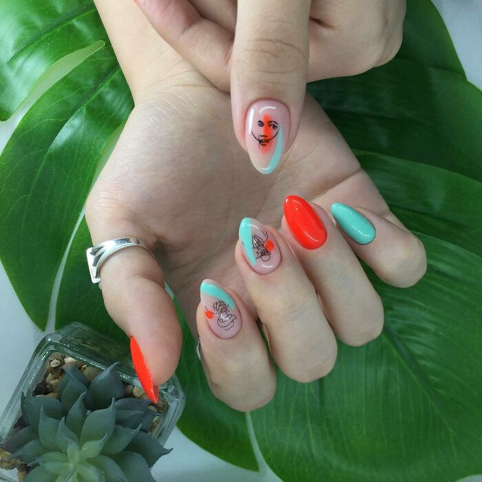 Hand with colorful nail art designs featuring abstract faces and bright blue and orange shades on green leaf background.