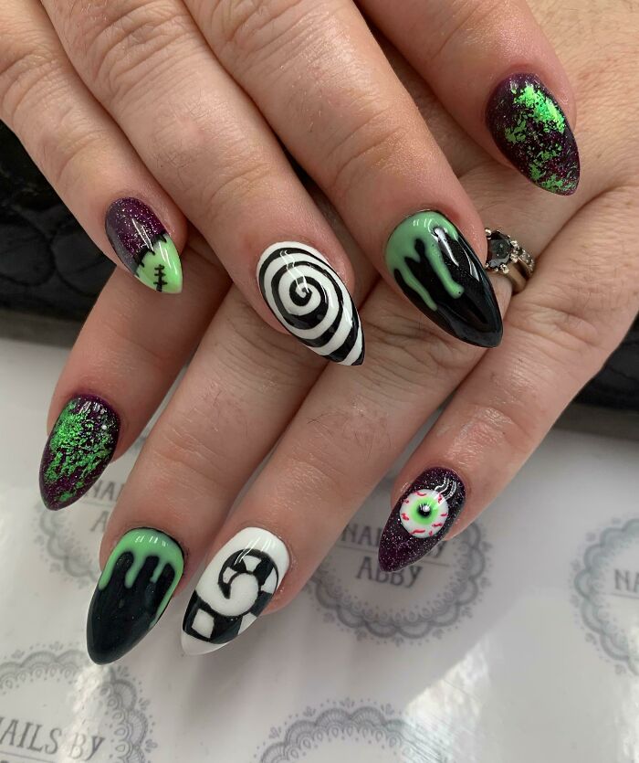 Halloween-inspired nail art with green, black, and purple designs featuring spirals, drips, and an eye motif.